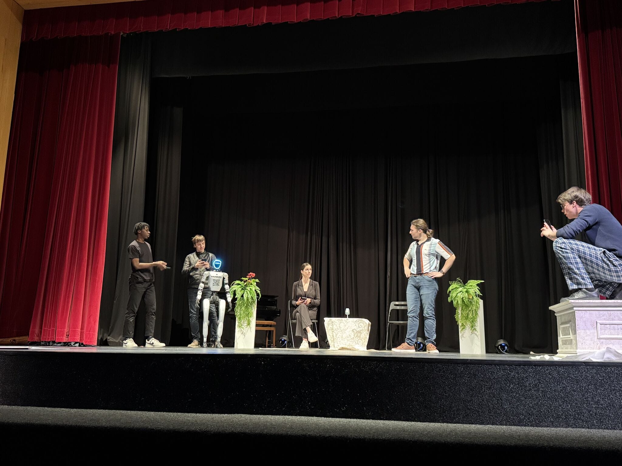 Wide shot of the theatre stage with the humanoid robot and the cast